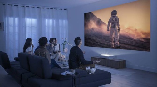 Grupo de amigos sentados no sofá da sala, assistindo a um filme de astronauta em um projetor. Estão de costas para a foto, são 3 homens e 2 mulheres. O sofá é preto e a sala é branca, o ambiente está com luz baixa.