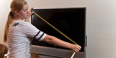 Uma mulher medindo o tamanho de uma TV com uma fita métrica. A TV está de costas para uma parede branca e a mulher, loira e de roupa branca, a mede de uma ponta a outra usando uma fita métrica amarela.
