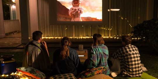 Um grupo de 4 pessoas sentadas na grama assistindo a um filme. É noite, o ambiente está iluminado com uma luz amarela baixa, as pessoas estão em almofadas e com roupas de frio.