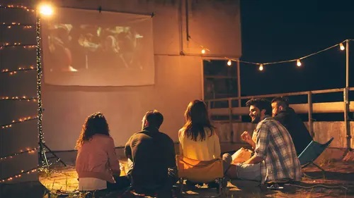 Um grupo de pessoas sentadas no chão assistindo a um filme na grama à noite, luzes amarelas em tom mais fraco. Cinco pessoas de costa, duas mulheres e três homens com uma projeção de um dos melhores projetores 4k ao fundo.